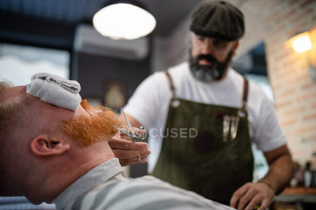 Barbier flou méconnaissable avec ciseaux fixant rousse homme barbe avec serviette couvrant les yeux assis dans le salon de coiffure — Photo de stock