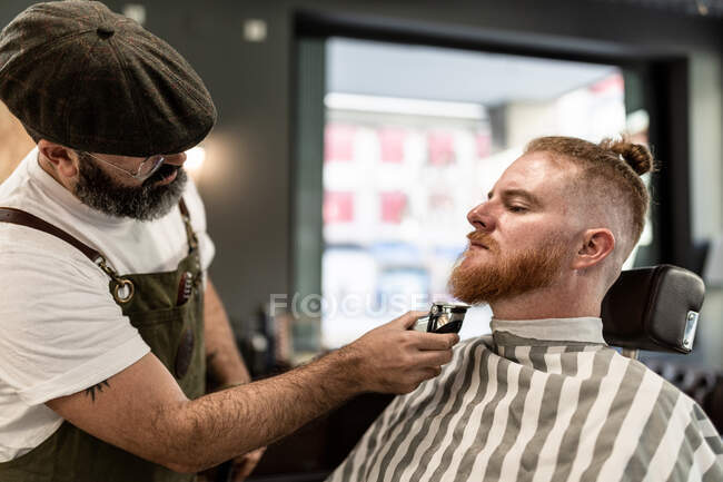 Barbeiro com pente e cortador de barba de corte de homem ruivo sentado na barbearia — Fotografia de Stock