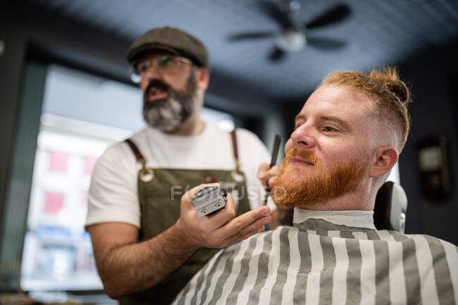 Barber with comb and trimmer cutting beard of redhead man sitting in barbershop — Stock Photo