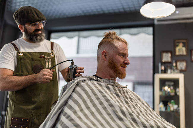 Barber applying fixative spray while making trendy hairstyle for confident relaxed man in cozy beauty salon — Stock Photo