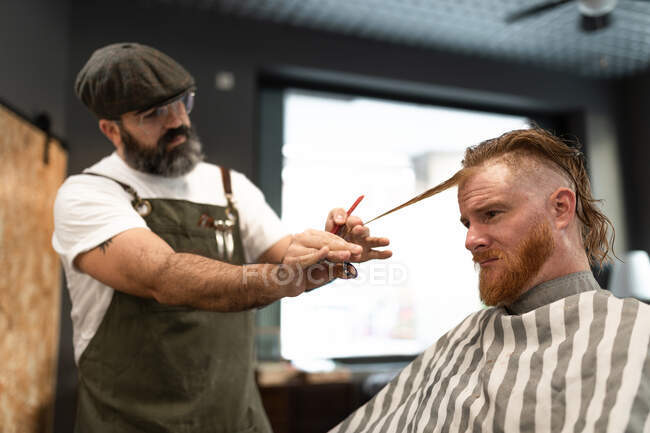 Modern hairdresser barber cutting a adult man redhead's hair in barber chair — Stock Photo