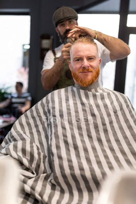 Сучасний перукар перукар, що ріже дорослий чоловік руде волосся в перукарському кріслі — стокове фото