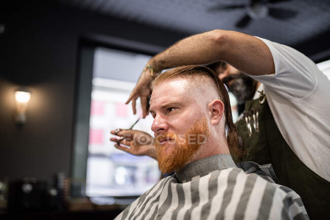 Cabeleireiro moderno barbeiro cortando o cabelo de um homem adulto ruiva em cadeira de barbeiro — Fotografia de Stock