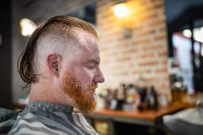 Вид сбоку на рыжего мужчину, сидящего в современной парикмахерской с закрытыми глазами в ожидании парикмахера — стоковое фото