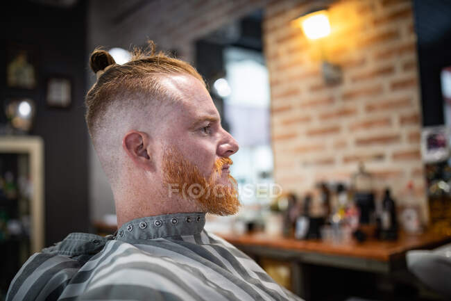 Vista lateral del pelirrojo sentado en la barbería moderna esperando a barbero - foto de stock