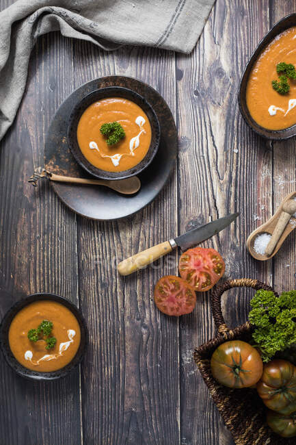 Вид сверху порций апельсинового томатного супа-пюре в стильных черных мисках на столе из лиственных пород с ломтиками помидоров — стоковое фото