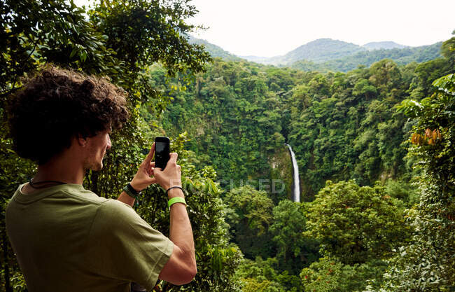 Back view of man with curly hair using smartphone to take photo of waterfall while travelling through green jungle in Costa Rica — Stock Photo