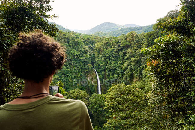 Неупізнаваний хлопець з кучерявим волоссям дивиться смартфон, стоячи навпроти водоспаду в зелених джунглях Коста-Рики. — стокове фото