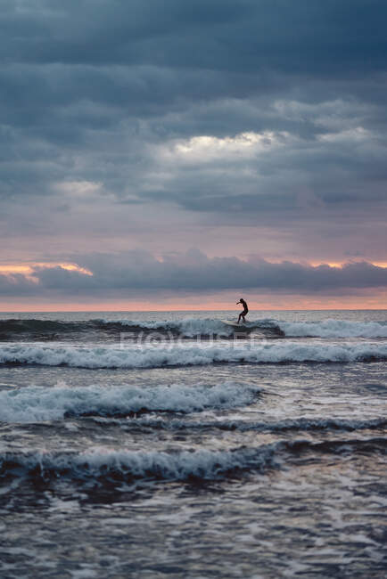Hombre irreconocible con tabla de surf caminando cerca del mar ondeando en la noche nublada en la playa en Costa Rica - foto de stock