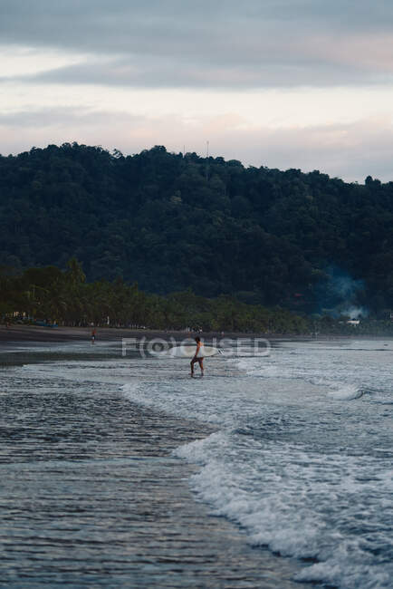 Uomo irriconoscibile con tavola da surf che cammina vicino al mare ondulato in serata nuvolosa sulla spiaggia in Costa Rica — Foto stock