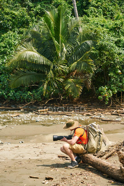 Side view of anonymous male tourist with backpack taking photos of landscape with professional camera while sitting in a tree branch on shore in Costa Rica — Stock Photo