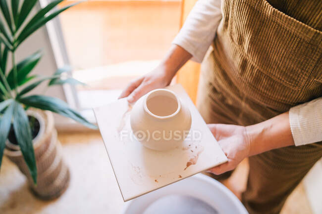 High angle of crop woman in casual clothes with small beige ceramic vase on square stand against blurred interior of light contemporary apartment — Stock Photo