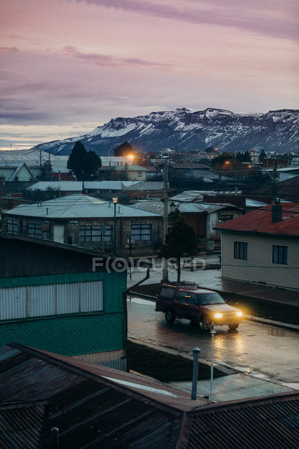 High angle of solitary pickup truck with luminous headlights on wet asphalt road near cottages and industrial buildings in town against snowy mountain ridge at horizon during sunset — Stock Photo