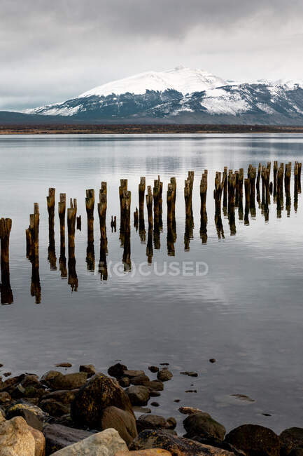 Pier de madeira destruído que conduz ao meio do lago calmo contra cume de montanha nevado — Fotografia de Stock