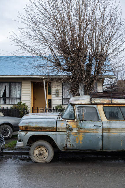 Velho automóvel abandonado cinza com pneu furado no quintal ao lado de árvore alta com ramos nus contra casa de campo em subúrbio sob céu nublado cinza — Fotografia de Stock