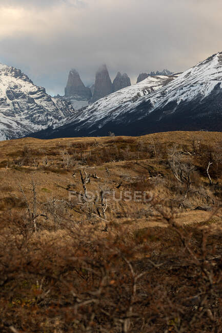 Meraviglioso scenario di colline con erba marrone secco contro cime innevate di montagne inclinate e rocce taglienti all'orizzonte in lussureggianti nuvole grigie — Foto stock