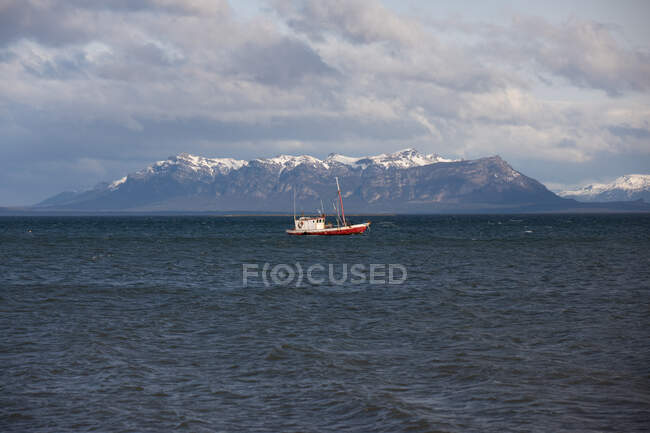 Одинокое судно в море среди водных волн против снежного горного хребта вдоль береговой линии в пасмурную погоду — стоковое фото