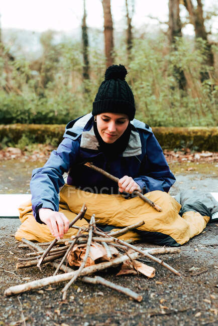 Fröhliche junge Frau in warmen Kleidern macht Lagerfeuer, während sie auf dem Boden sitzt, während sie im Herbst auf dem Land campt — Stockfoto