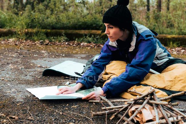 Mochilera joven concentrada que busca ubicación en el mapa mientras está sentada en el suelo durante el trekking en el bosque en el día de otoño - foto de stock