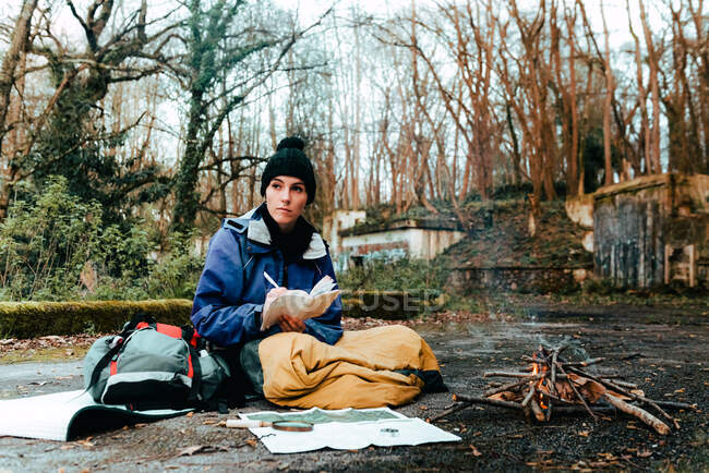 Senderista joven reflexiva escribiendo información sobre la ruta de trekking mientras descansa en la hoguera en el campo en el día de otoño - foto de stock