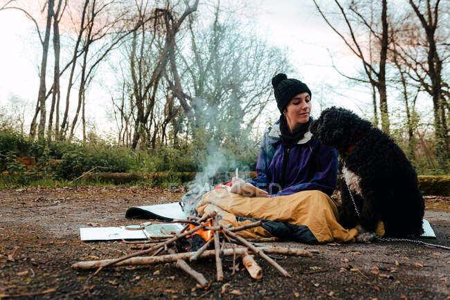 Zufriedene junge Wanderin sieht schwarzen Hund beim gemeinsamen Aufwärmen am Lagerfeuer im Wald an einem Herbsttag — Stockfoto
