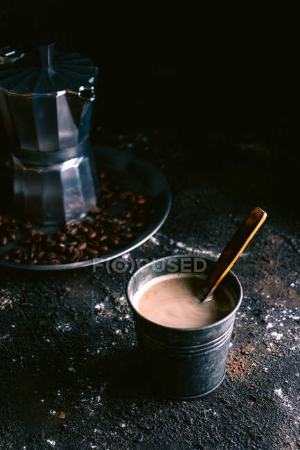 Copo de metal com café acabado de fazer e colher de madeira colocada na superfície preta bagunçada perto da bandeja com cafeteira e grãos torrados — Fotografia de Stock