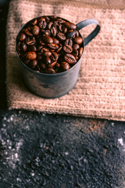 De arriba taza de metal con granos de café tostados colocados en la servilleta de tela en la mesa áspera - foto de stock