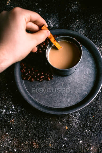 Зверху анонімний чоловік перемішує свіжу каву з дерев'яною ложкою в металевій чашці на підносі зі смаженими бобами над брудним столом — стокове фото