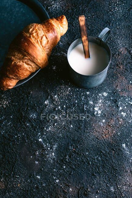 De acima mencionado pão fresco e caneca de leite com colher colocada na superfície preta suja durante o café da manhã — Fotografia de Stock