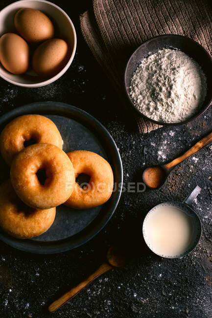 Вид зверху на свіжі пончики, розміщені на грубому столі біля різних інгредієнтів випічки та посуду на кухні — стокове фото
