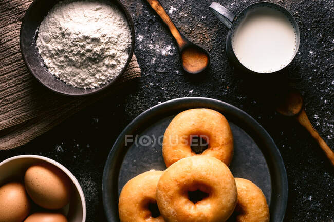 Vista superior de donuts frescos colocados em mesa áspera perto de vários ingredientes de pastelaria e utensílios na cozinha — Fotografia de Stock