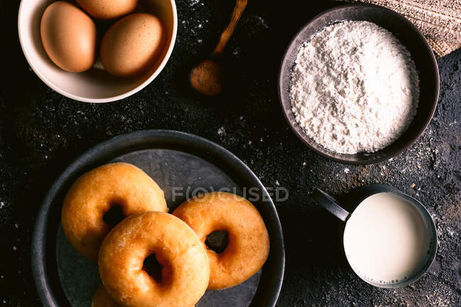 Vista dall'alto di ciambelle fresche poste su tavola ruvida vicino a vari ingredienti e utensili da pasticceria in cucina — Foto stock