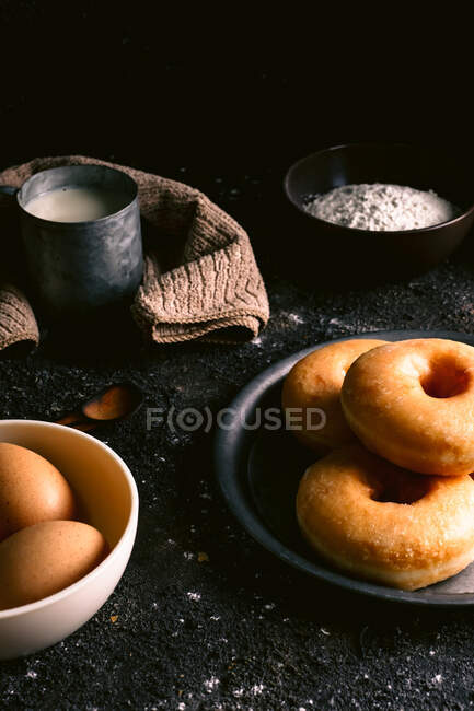 Ciambelle fresche poste su tavola ruvida vicino a vari ingredienti e utensili da pasticceria in cucina — Foto stock