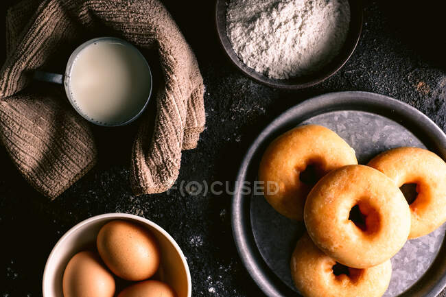 Top view of fresh donghnuts placed on rough table near various pastry ingredients and utensils in kitchen — стоковое фото