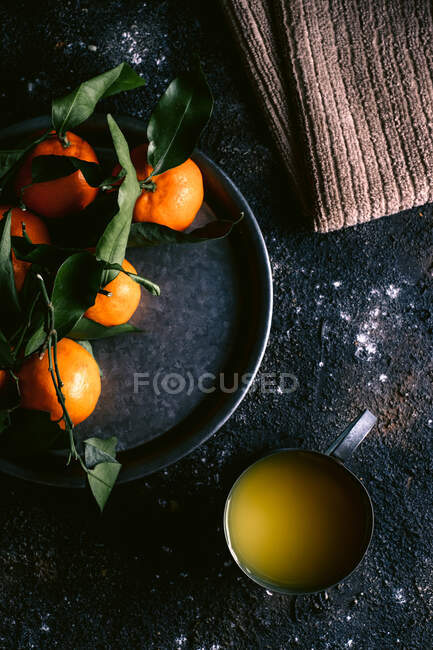 Vista superior de la taza con jugo de fruta fresca y plato con mandarinas maduras colocadas en la mesa negra áspera cerca de la servilleta - foto de stock