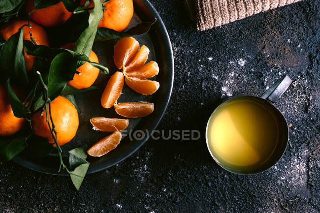 Top view of mug with fresh fruit juice and plate with ripe tangerines placed on rough black table near napkin — Stock Photo