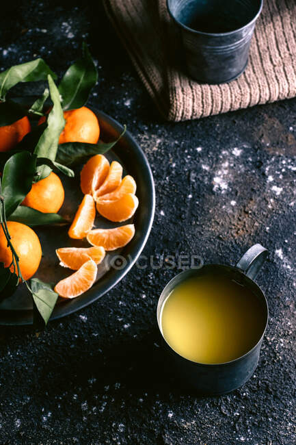 Vue du dessus de la tasse avec jus de fruits frais et assiette avec mandarines mûres placées sur une table noire rugueuse près de la serviette — Photo de stock