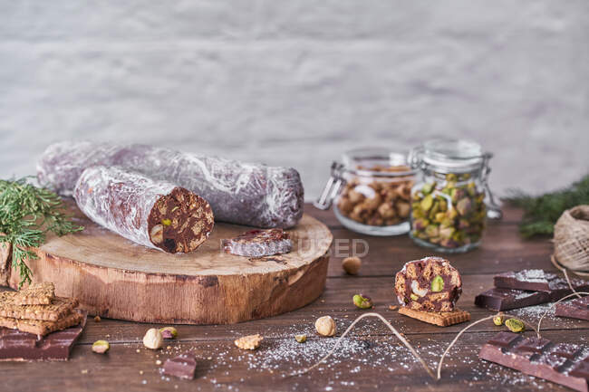 Traditional homemade chocolate salami with hazelnuts and pistachios on wooden board placed on table with ingredients and green spruce branches — Stock Photo