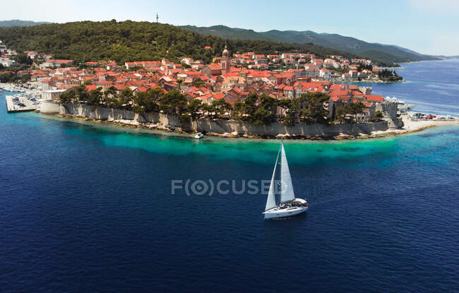 Краєвид на старе містечко на острові з зеленими тропічними рослинами і білим вітрильником серед блакитного спокійного океану в Хорватії. — стокове фото