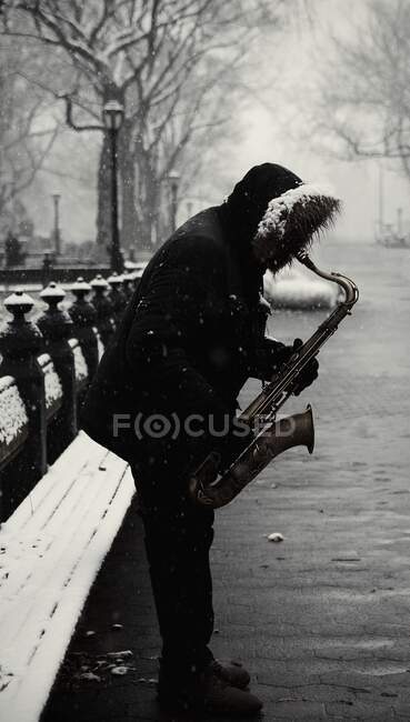 Vista lateral do homem sem rosto em casaco preto quente com capuz de pé e tocando saxofone durante o inverno frio em Nova York — Fotografia de Stock