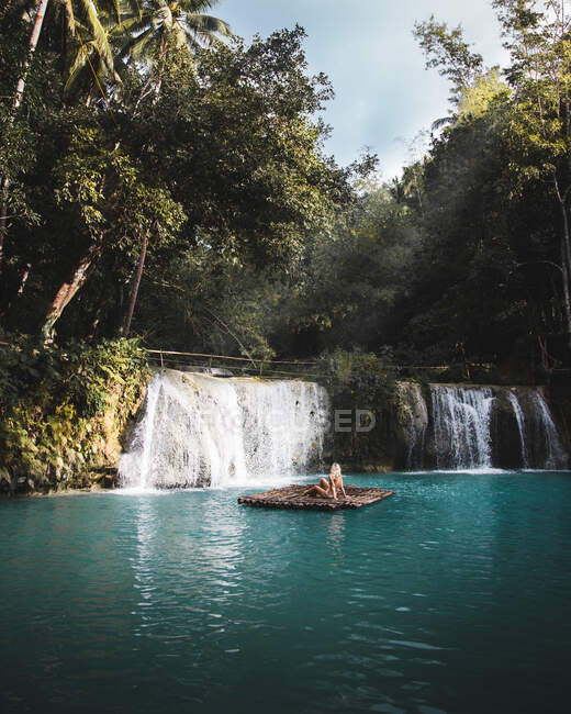 Anonymous woman on wooden raft among turquoise water near small waterfall in tropical green jungle — Stock Photo