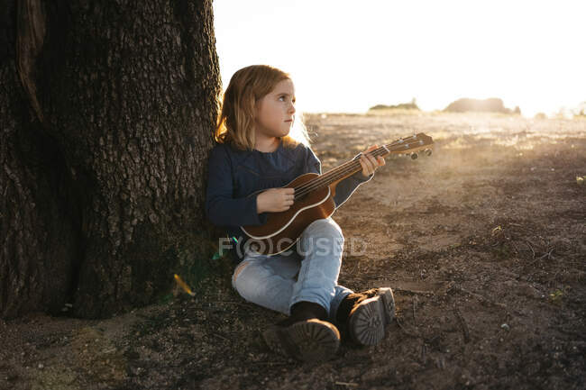 Adorabile bambina seria in abbigliamento casual suonare la chitarra ukulele mentre seduto vicino all'albero in soleggiata giornata estiva in campagna — Foto stock