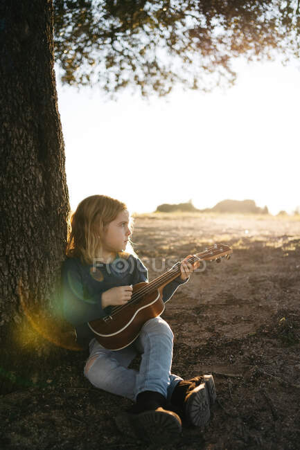 Очаровательная серьезная девочка в повседневной одежде играет на гитаре укулеле, сидя рядом с деревом в солнечный летний день в сельской местности — стоковое фото