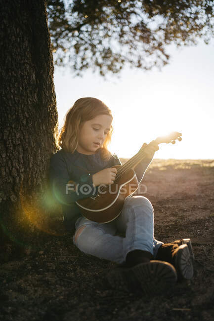 Adorable serious little girl in casual wear playing ukulele guitar while sitting near tree in sunny summer day in countryside — Stock Photo