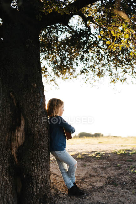 Adorable niña seria en ropa casual tocando la guitarra ukelele mientras está de pie cerca del árbol en el soleado día de verano en el campo - foto de stock