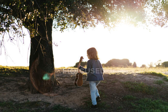 Обратный вид на неузнаваемую маленькую девочку в повседневной одежде идущую с гитарой укулеле рядом с деревом в солнечный летний день в сельской местности — стоковое фото