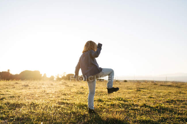 Back view of anonymous adorable active little girl in casual wear playing and dancing in green field while enjoying sunny summer evening in countryside — Stock Photo