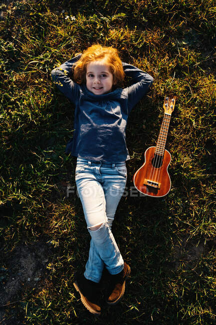 Сверху очаровательная маленькая девочка лежит на траве и улыбается, глядя в камеру возле гитары укулеле в солнечный летний день в сельской местности. — стоковое фото