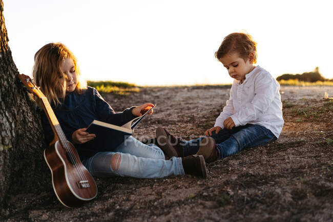 Vue latérale d'une adorable petite fille lisant une histoire intéressante pour son jeune frère assis sous un arbre avec une guitare ukulélé en été à la campagne — Photo de stock