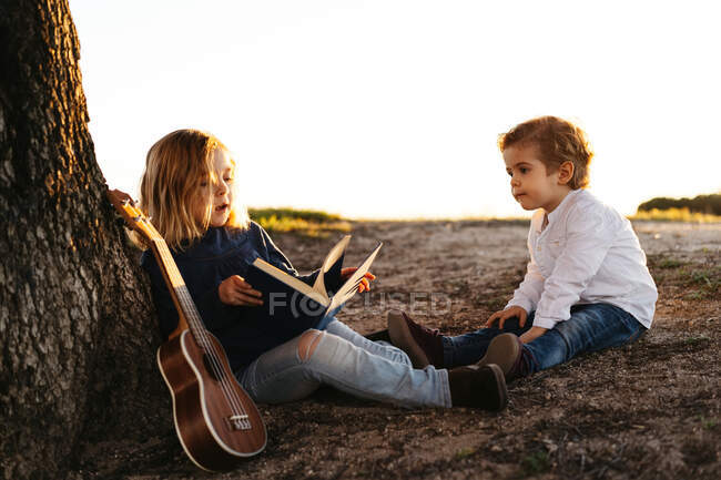 Vista lateral de la adorable niña leyendo interesante historia a su hermano menor mientras se sientan juntos bajo el árbol con la guitarra ukelele en el día de verano en el campo - foto de stock